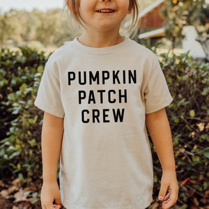 May include: A child wearing a beige t-shirt with the text "Pumpkin Patch Crew" printed in black.