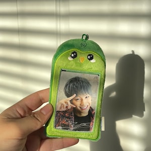 May include: A green avocado-shaped card holder with a photo of a person inside. The card holder has a cartoon face with black eyes and a yellow beak. The photo shows a person with gray hair and a red and black patterned shirt.