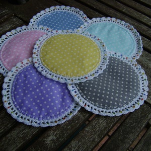 Puede incluir: Seis posavasos de ganchillo con borde blanco y centros de tela con lunares de colores. Los posavasos están dispuestos en círculo sobre una superficie de madera.