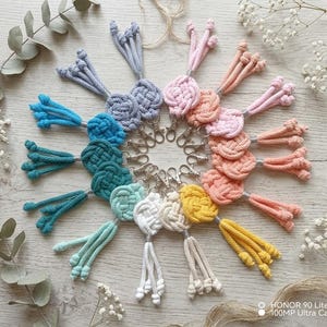 May include: A selection of colourful macrame keyrings, featuring knotted designs and tassels. The keyrings are in shades of blue, green, pink, and yellow, each attached to a silver-coloured clasp. The keyrings are arranged in a circular pattern on a light wooden surface.
