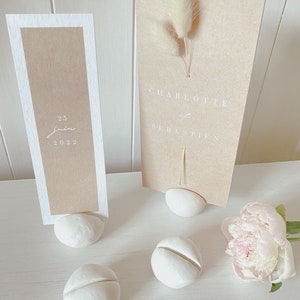 May include: Four white ceramic table number holders with a groove on the top. Two holders have cards with text, one says "25 juin 2022" and the other says "Charlotte & Sebastien".
