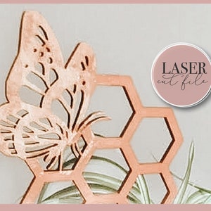 Op de afbeelding: Een lasergesneden ontwerp met een honingraatpatroon met een vlinder erop. Het ontwerp is gemaakt van hout en is geverfd in een lichtbruine kleur. De tekst "LASER cut file" is zichtbaar in een cirkel aan de rechterkant van de afbeelding.