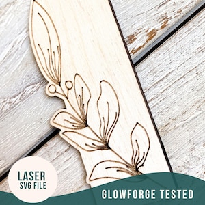 May include: A close-up of a wooden bookmark with a laser-cut floral design. The bookmark is light brown and the design is in a dark brown color. The text "LASER SVG FILE" is visible in a circle on the left side of the image. The text "GLOWFORGE TESTED" is visible in the bottom right corner of the image.