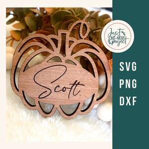 May include: A wooden pumpkin-shaped cutout with the name "Scott" engraved in a cursive font. The pumpkin is brown with a decorative cutout design. The image also includes the text "SVG, PNG, DXF" and a logo that says "Just One More Project".