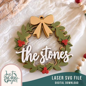 Op de afbeelding: Een gelaagd houten ornament in de vorm van een krans met groene hulstbladeren, rode bessen en een beige strik aan de bovenkant. In het midden staat de witte tekst "the stones". Het ontwerp heeft een klein gaatje om het op te hangen.