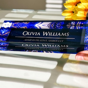 May include: A blue and gold glass desk nameplate with the text "Olivia Williams, Administrative Assistant" engraved on it.