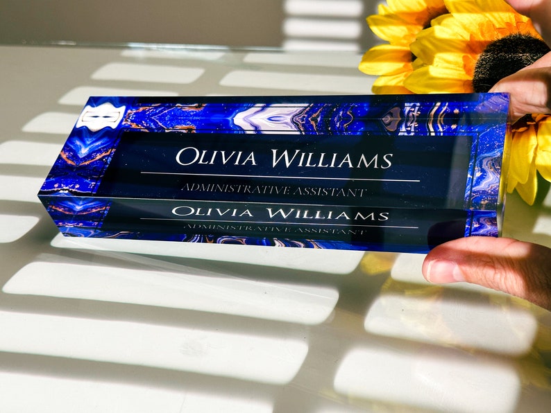 May include: A blue and gold glass desk nameplate with the text "Olivia Williams, Administrative Assistant" engraved on it.