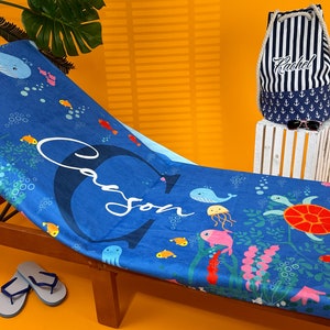May include: A blue beach towel with a sea life design, including fish, a whale, an octopus, a jellyfish, and a turtle. The towel has the name "Cason" written on it in white cursive lettering. The towel is lying on a wooden lounge chair with a pair of blue flip flops on the ground next to it.