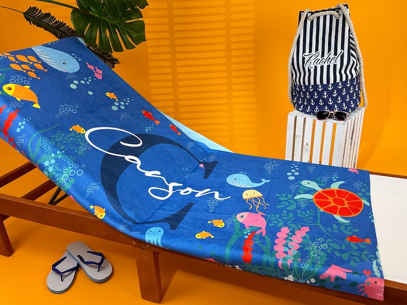 May include: A blue beach towel with a sea life design, including fish, a whale, an octopus, a jellyfish, and a turtle. The towel has the name "Cason" written on it in white cursive lettering. The towel is lying on a wooden lounge chair with a pair of blue flip flops on the ground next to it.
