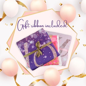 May include: A gift-wrapped set of three purple, pink, and white blankets with glitter accents. The blankets are tied with a brown ribbon and the text "Gift ribbon included" is written in purple on a pink background.