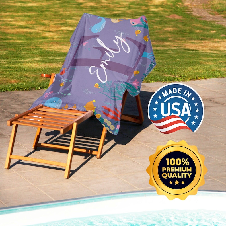 May include: A purple beach towel with a sea life design and the name "Emily" written in white. The towel is folded and placed on a wooden chair. The chair is next to a swimming pool. There are two labels on the image. One label says "Made in USA" and the other says "100% Premium Quality".