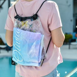 May include: A clear plastic drawstring backpack with a blue and white ombre beach towel inside. The towel has the name "Olivia" printed on it in white.