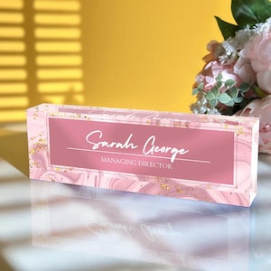May include: Pink acrylic desk nameplate with gold glitter accents. The nameplate features the text "Sarah George, Managing Director".