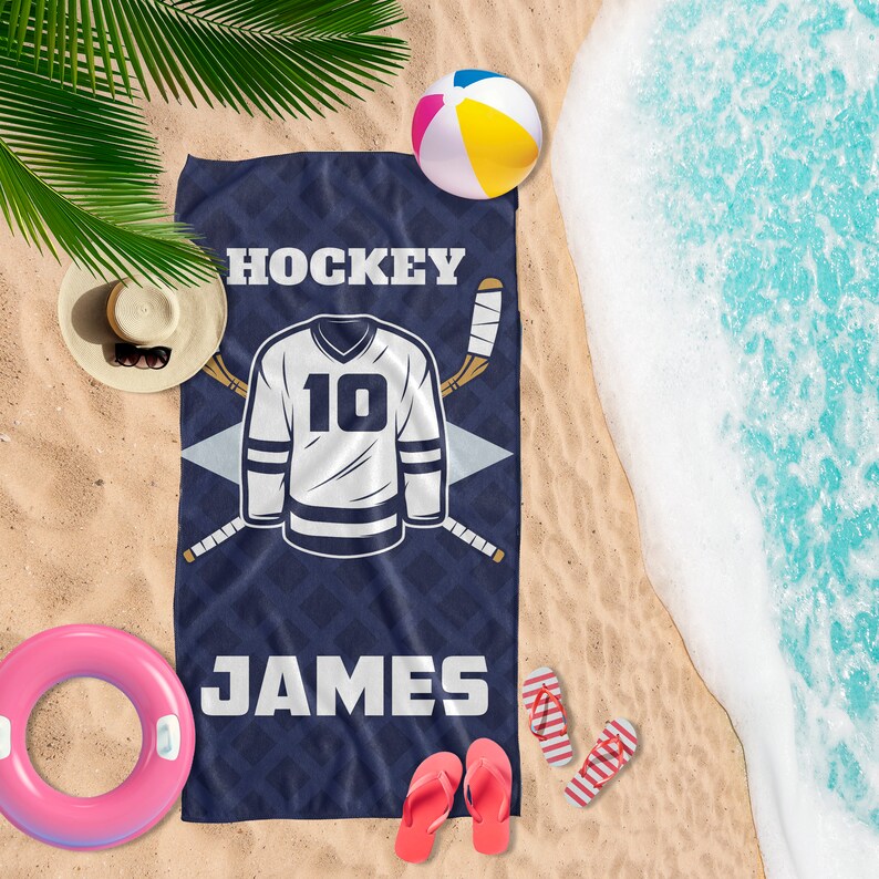 May include: Navy blue beach towel with a hockey jersey design. The towel features the word "HOCKEY" at the top, a white jersey with the number 10, and the name "JAMES" at the bottom. Two hockey sticks are crossed behind the jersey.