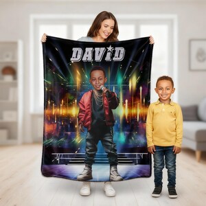 May include: A black blanket featuring a cartoon illustration of a young boy holding a microphone on a stage, with the name "DAVID" in white letters. The boy is wearing a red jacket and black pants. A woman and a young boy are standing in front of the blanket.