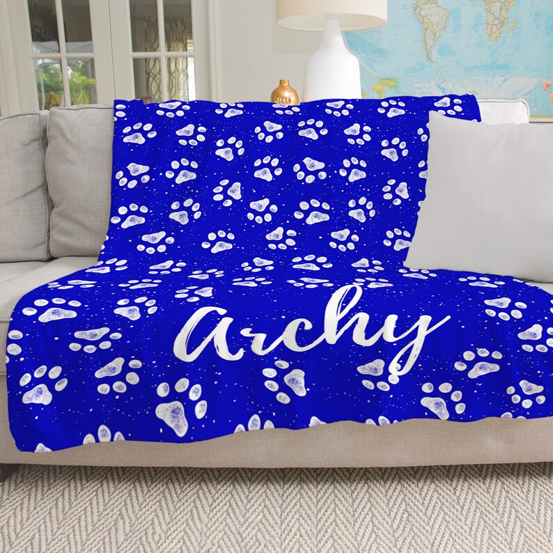 May include: A blue fleece throw blanket with a white paw print pattern and the name "Archie" written in white cursive lettering.