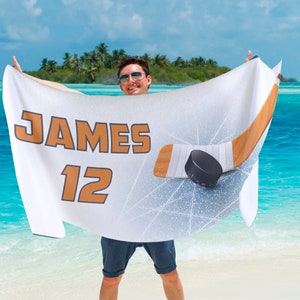 May include: White hockey-themed beach towel with the name "JAMES" and the number "12" in orange. A hockey stick and puck graphic are displayed. The towel is held up against a tropical beach backdrop.