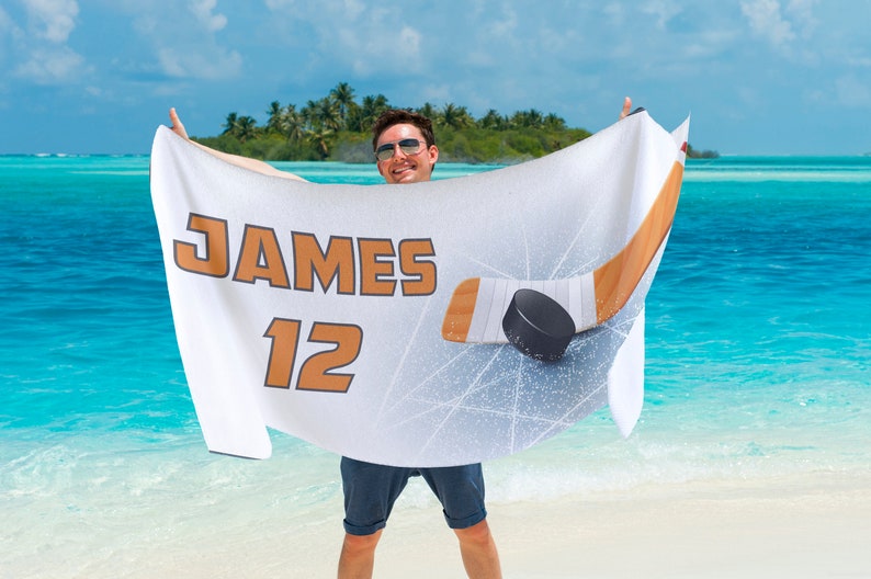 May include: White hockey-themed beach towel with the name "JAMES" and the number "12" in orange. A hockey stick and puck graphic are displayed. The towel is held up against a tropical beach backdrop.