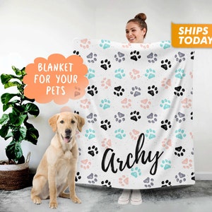 May include: White fleece blanket with a paw print pattern in black, pink, and blue. The blanket has the name "Archie" written in black cursive lettering.