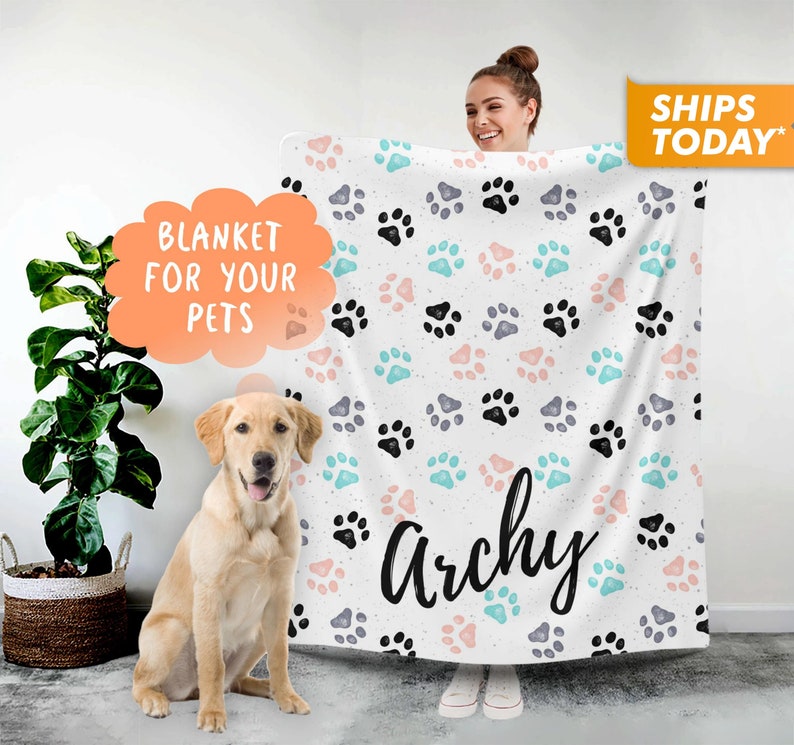 May include: White fleece blanket with a paw print pattern in black, pink, and blue. The blanket has the name "Archie" written in black cursive lettering.
