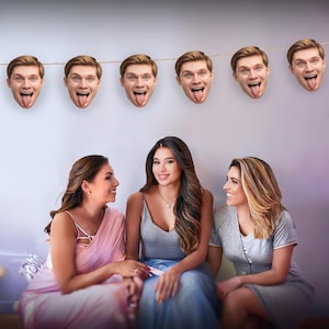 May include: A decorative garland of smiling male faces with tongues sticking out hangs above three women. The women are smiling and sitting together. One woman wears a pink dress, another a gray top and blue skirt, and the third a gray dress.