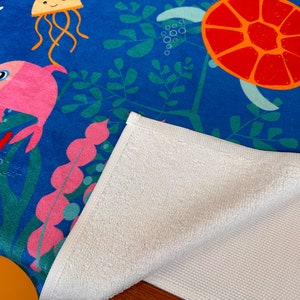May include: A blue beach towel with a colorful underwater scene featuring a pink fish, a jellyfish, and a sea turtle. The towel has a white terry cloth backing.