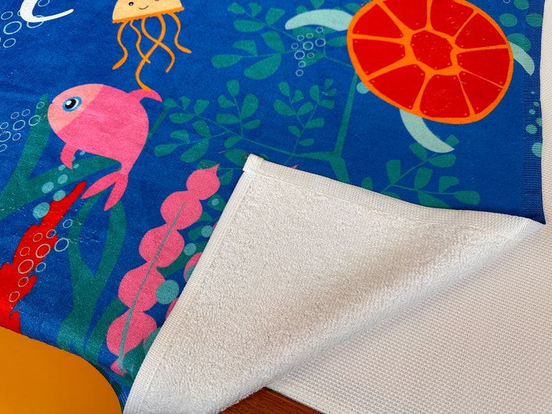 May include: A blue beach towel with a colorful underwater scene featuring a pink fish, a jellyfish, and a sea turtle. The towel has a white terry cloth backing.