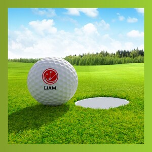 May include: A white golf ball with a red logo and the name "LIAM" printed on it. The ball is on a green grassy surface near a golf hole.