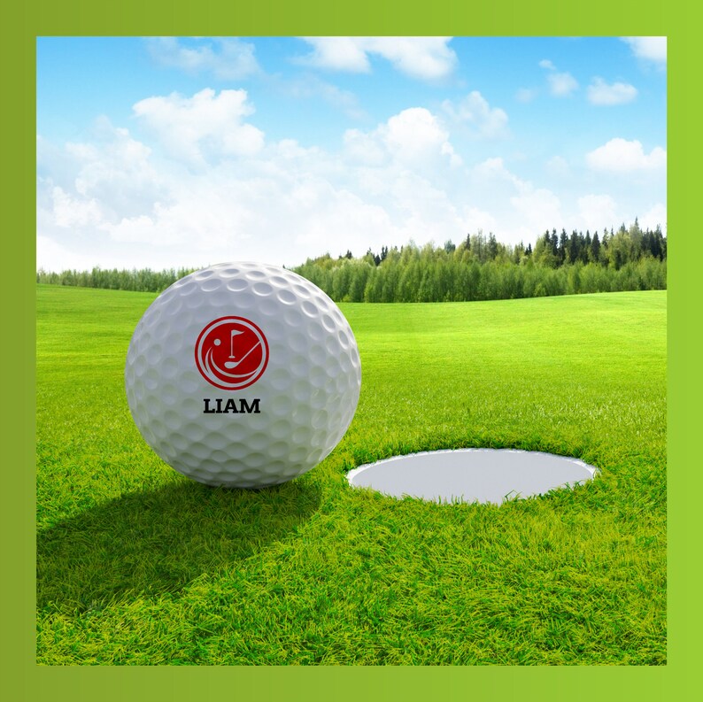 May include: A white golf ball with a red logo and the name "LIAM" printed on it. The ball is on a green grassy surface near a golf hole.