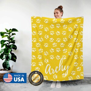 May include: A yellow fleece blanket with a white paw print pattern. The blanket has the name "Archie" written in white text on it.