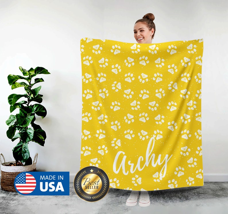 May include: A yellow fleece blanket with a white paw print pattern. The blanket has the name "Archie" written in white text on it.