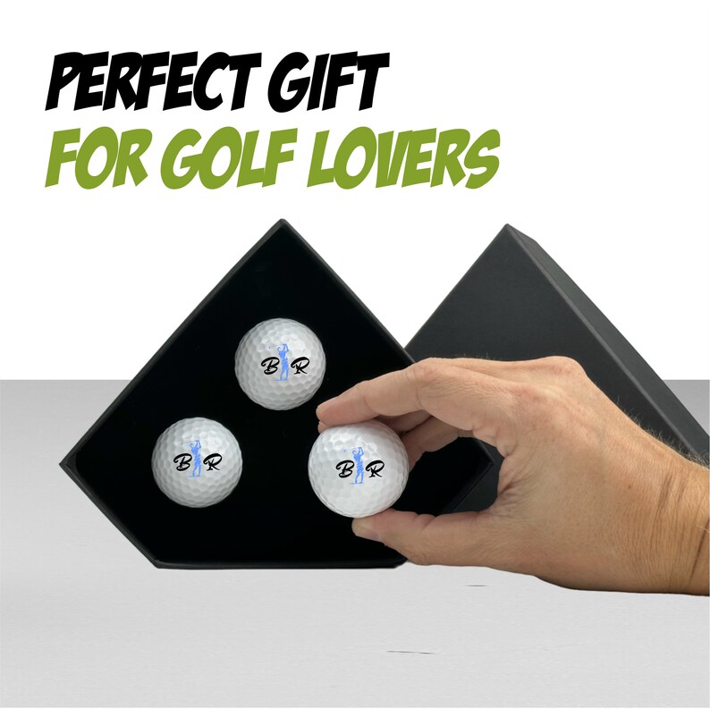 May include: Three white golf balls with a blue golfer graphic and the letters "B.R." printed on them are displayed in a black gift box.