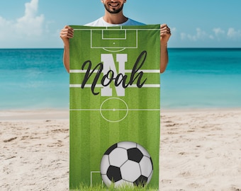 May include: Green soccer field beach towel with the name "Noah" in white script. The towel features a soccer ball at the bottom and field markings. The background is a beach with blue water and sky.