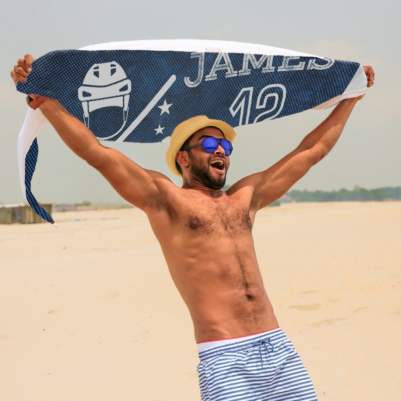 May include: A person on a beach holds up a blue and white towel with the name "JAMES" and the number "19". The towel features a hockey helmet graphic and stars. The person wears a straw hat, sunglasses, and blue and white striped swim shorts.