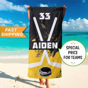 May include: Black and yellow hockey towel with the name "Aiden" and the number "33". The towel features a hockey puck and crossed hockey sticks in white. The background is a beach scene with blue water and sky.