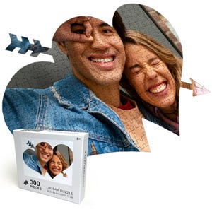 May include: A 300-piece jigsaw puzzle featuring a couple smiling and looking at the camera. The puzzle is shaped like a heart with an arrow through it. The box is white with a picture of the puzzle on the front and the text "300 PIECES JIGSAW PUZZLE 58.5x42cm 23x16.5in 8+"