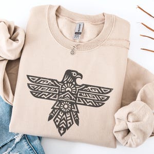 May include: A tan sweatshirt with a black graphic of an eagle with geometric patterns. The sweatshirt is folded with a silver necklace and a pair of blue jeans. The Gildan brand label is visible.