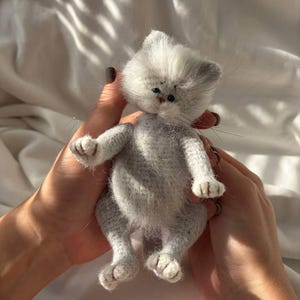 May include: A small, fuzzy, light grey kitten figurine crafted with a textured, yarn-like appearance. The kitten has a rounded head, pointed ears, dark eyes, a small nose, and long, thin whiskers extending from its face and paws.