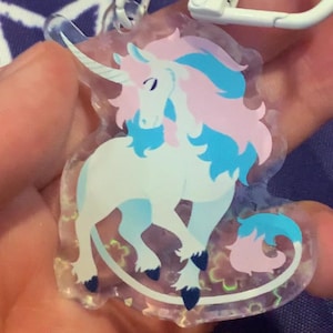 May include: A clear acrylic keychain featuring a cartoon unicorn with a white body, pink and blue mane and tail, and a white horn. The keychain has a white metal clasp.
