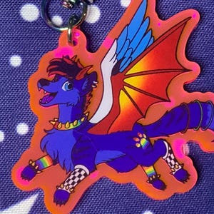 May include: A colorful keychain featuring a cartoon wolf with wings. The wolf is blue with a red and orange wing design, rainbow accents, and a gold collar. The keychain has a silver clasp and is set against a blue background.