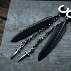 Może przedstawiać: Para czarnych piórkowych kolczyków z srebrnymi łańcuszkami i zawieszkami w kształcie sztyletu. Kolczyki są wykonane z metalu i mają czarne wykończenie.