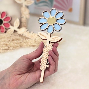 May include: A wooden flower-shaped keepsake with a light blue and gold center. The flower has a stem with the word "Parrain" and the name "Sandro" and date "04-05-2025" engraved on it. The word "Bapt&ecirc;me" is also engraved on the flower.