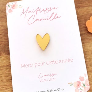 May include: A gold heart-shaped pin on a white card with pink floral accents. The card has text in French, including "Ma&icirc;tresse Camille" and "Merci pour cette ann&eacute;e". A small orange flower-shaped object is visible.