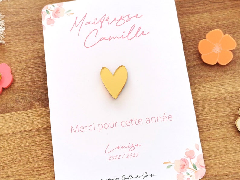 May include: A gold heart-shaped pin on a white card with pink floral accents. The card has text in French, including "Ma&icirc;tresse Camille" and "Merci pour cette ann&eacute;e". A small orange flower-shaped object is visible.