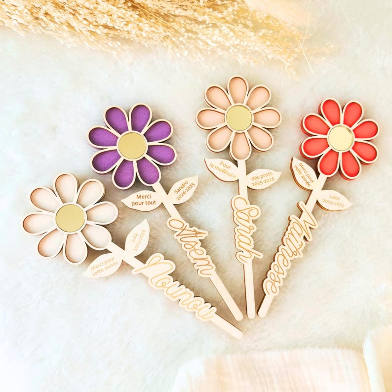 May include: Wooden flower-shaped decorations with colorful petals and gold centers. Each flower has a stem with a name and text like "Merci pour tout" and "Sandro 2024-2025".
