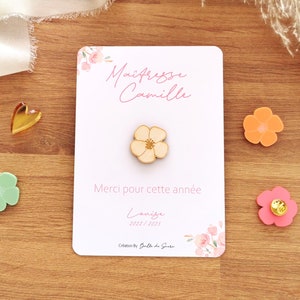 May include: A wooden flower pin on a white card with the text "Ma&icirc;tresse Camille" and "Merci pour cette ann&eacute;e". The card is decorated with floral accents. Other flower-shaped pins are scattered around.