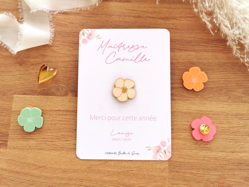 May include: A wooden flower pin on a white card with the text "Ma&icirc;tresse Camille" and "Merci pour cette ann&eacute;e". The card is decorated with floral accents. Other flower-shaped pins are scattered around.