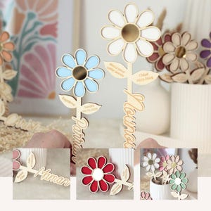 May include: Wooden flower-shaped decorations with personalized text. The flowers have petals in various colors, including blue, white, and red, with a gold center. Some flowers have names and dates engraved on them.