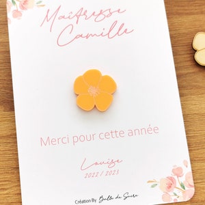 May include: An orange flower-shaped pin is centered on a white card with the text "Ma&icirc;tresse Camille" and "Merci pour cette ann&eacute;e" in pink. A wooden flower pin and a pink flower pin are also visible.