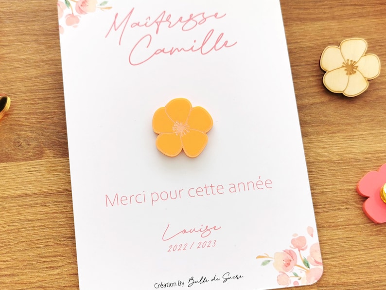 May include: An orange flower-shaped pin is centered on a white card with the text "Ma&icirc;tresse Camille" and "Merci pour cette ann&eacute;e" in pink. A wooden flower pin and a pink flower pin are also visible.